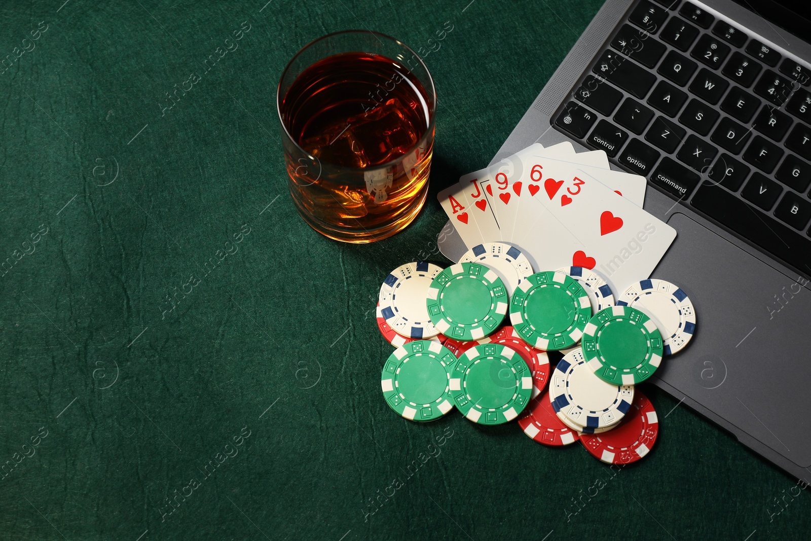 Photo of Poker chips, laptop, playing cards and glass of alcohol drink on green table, flat lay. Online game