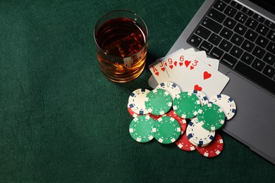 Photo of Poker chips, laptop, playing cards and glass of alcohol drink on green table, flat lay. Online game