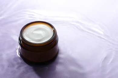 Photo of Jar with face cream in water on light violet background, space for text