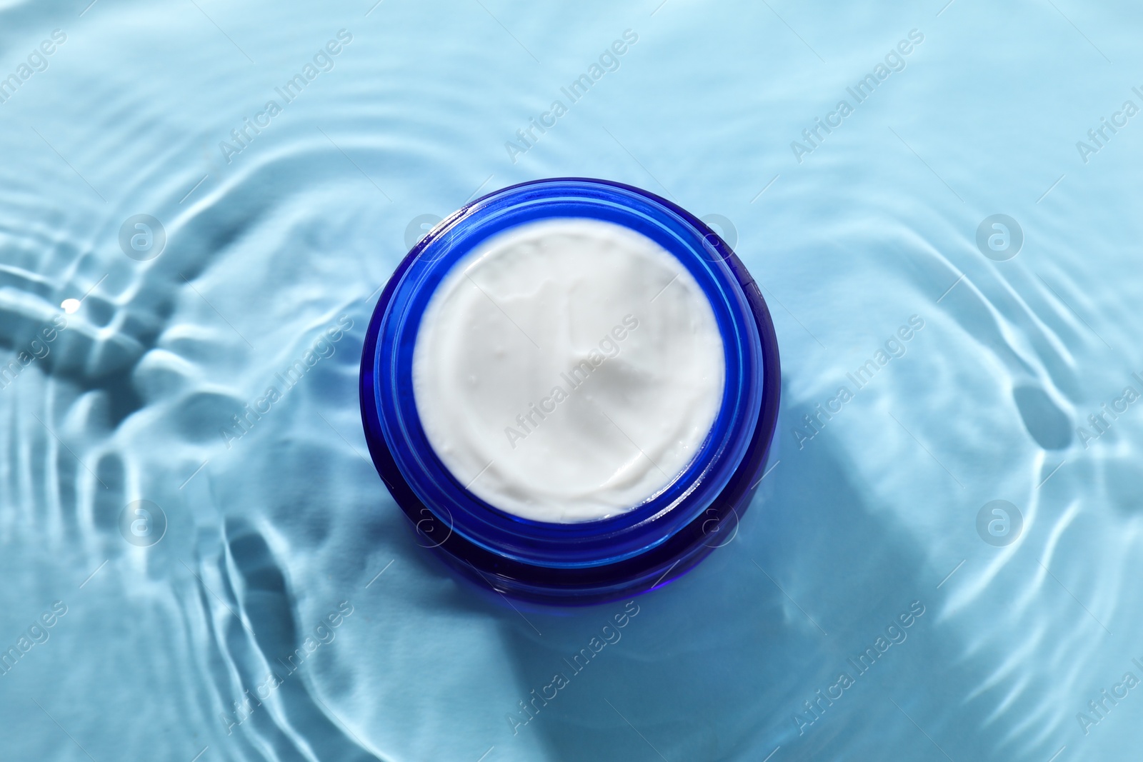 Photo of Jar with face cream in water on light blue background, top view