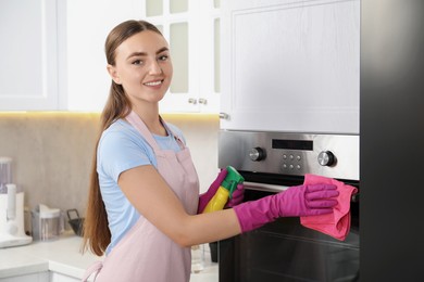 Photo of Young woman wiping electric oven with rag and bottle of detergent in kitchen