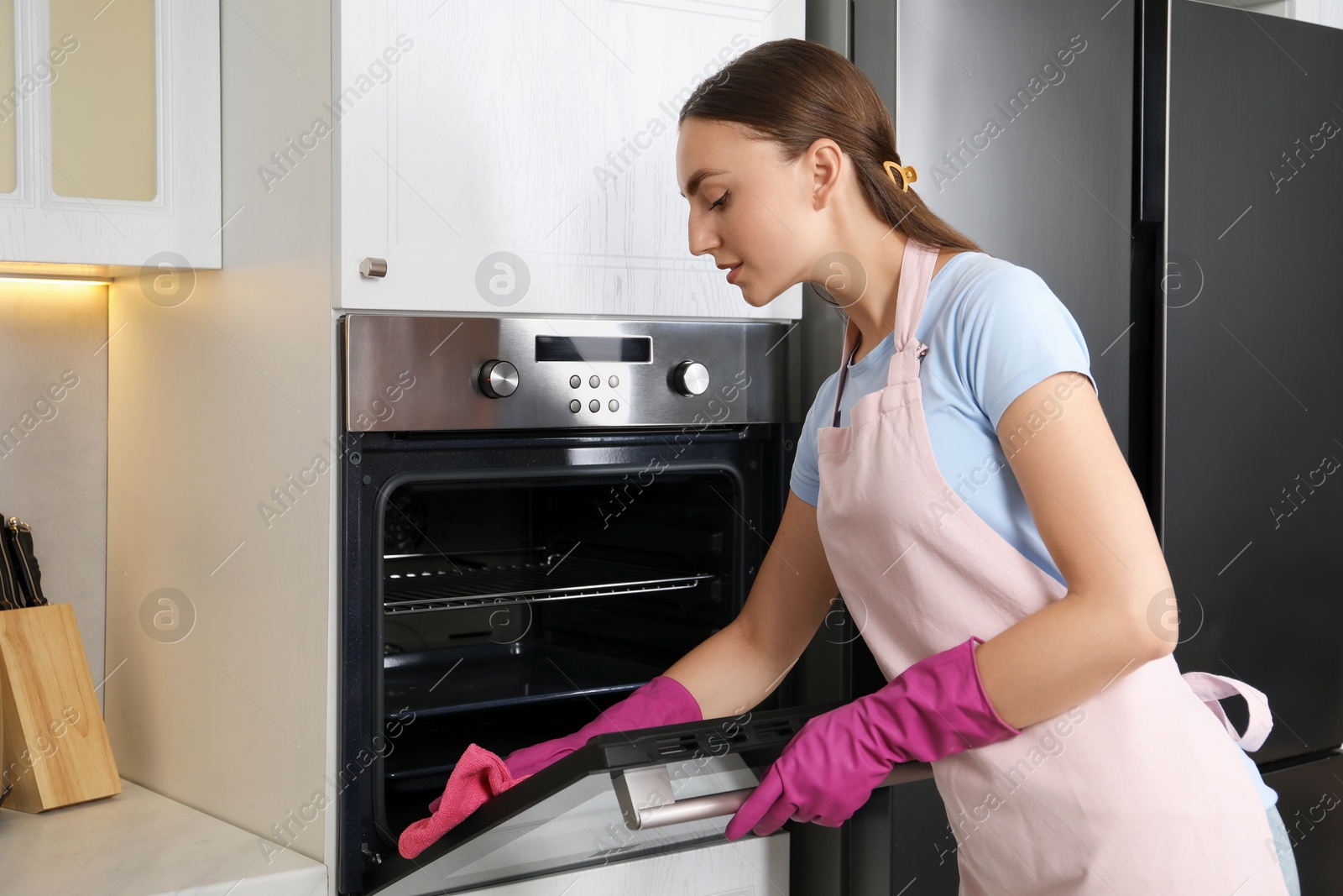 Photo of Beautiful young woman wiping electric oven with rag in kitchen