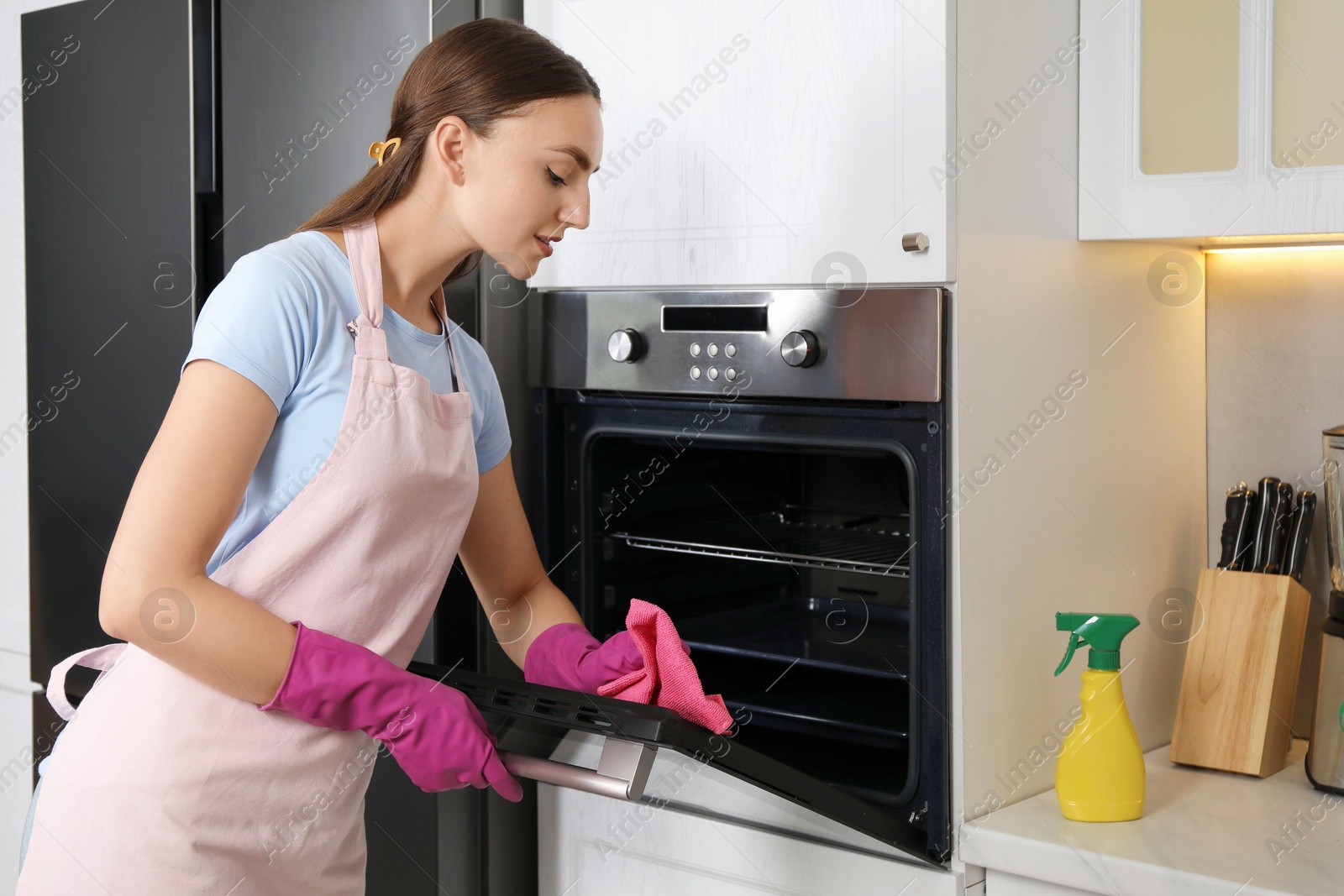 Photo of Beautiful young woman wiping electric oven with rag in kitchen