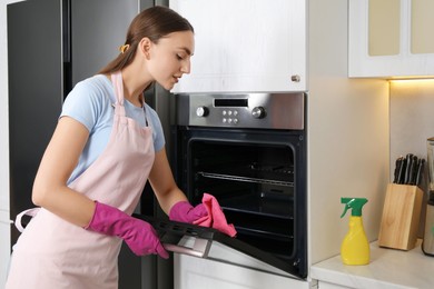 Photo of Beautiful young woman wiping electric oven with rag in kitchen