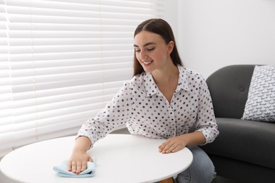 Photo of Beautiful young woman wiping coffee table with rag at home