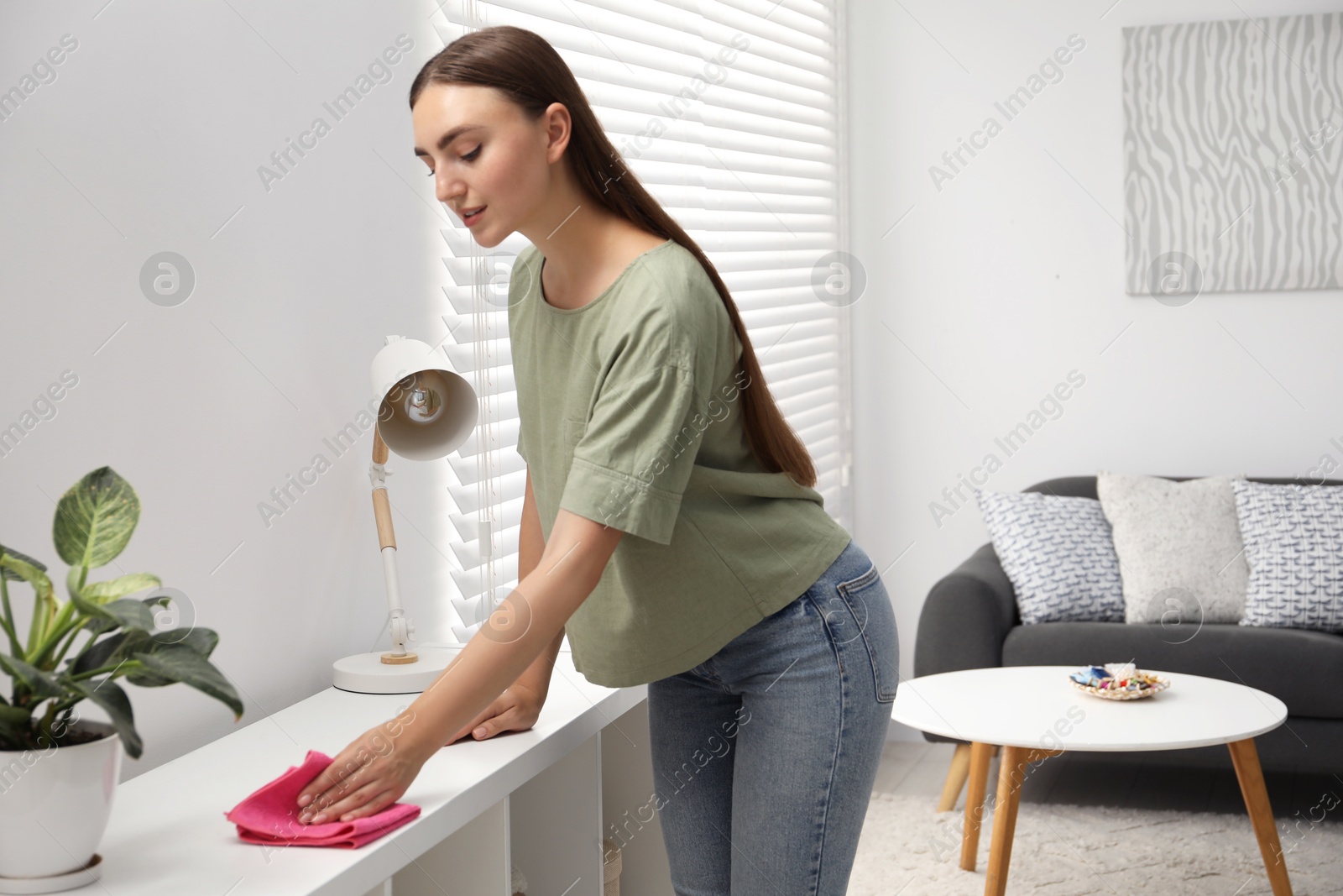 Photo of Beautiful young woman wiping furniture with rag at home