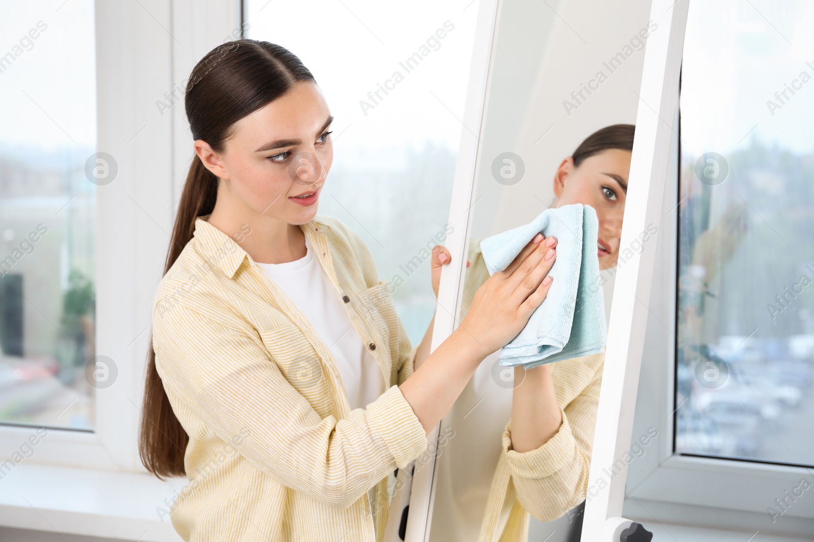 Photo of Beautiful young woman cleaning mirror with rag at home