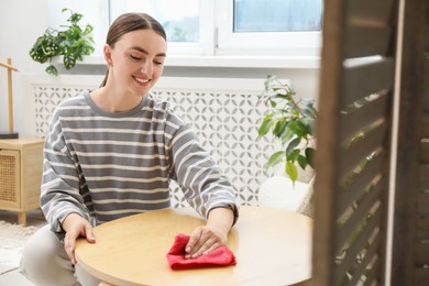 Photo of Beautiful young woman wiping coffee table with rag at home