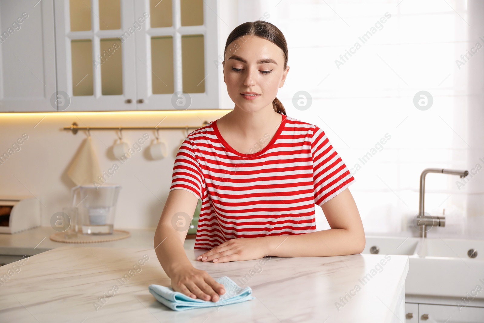 Photo of Beautiful young woman cleaning white marble table with rag in kitchen