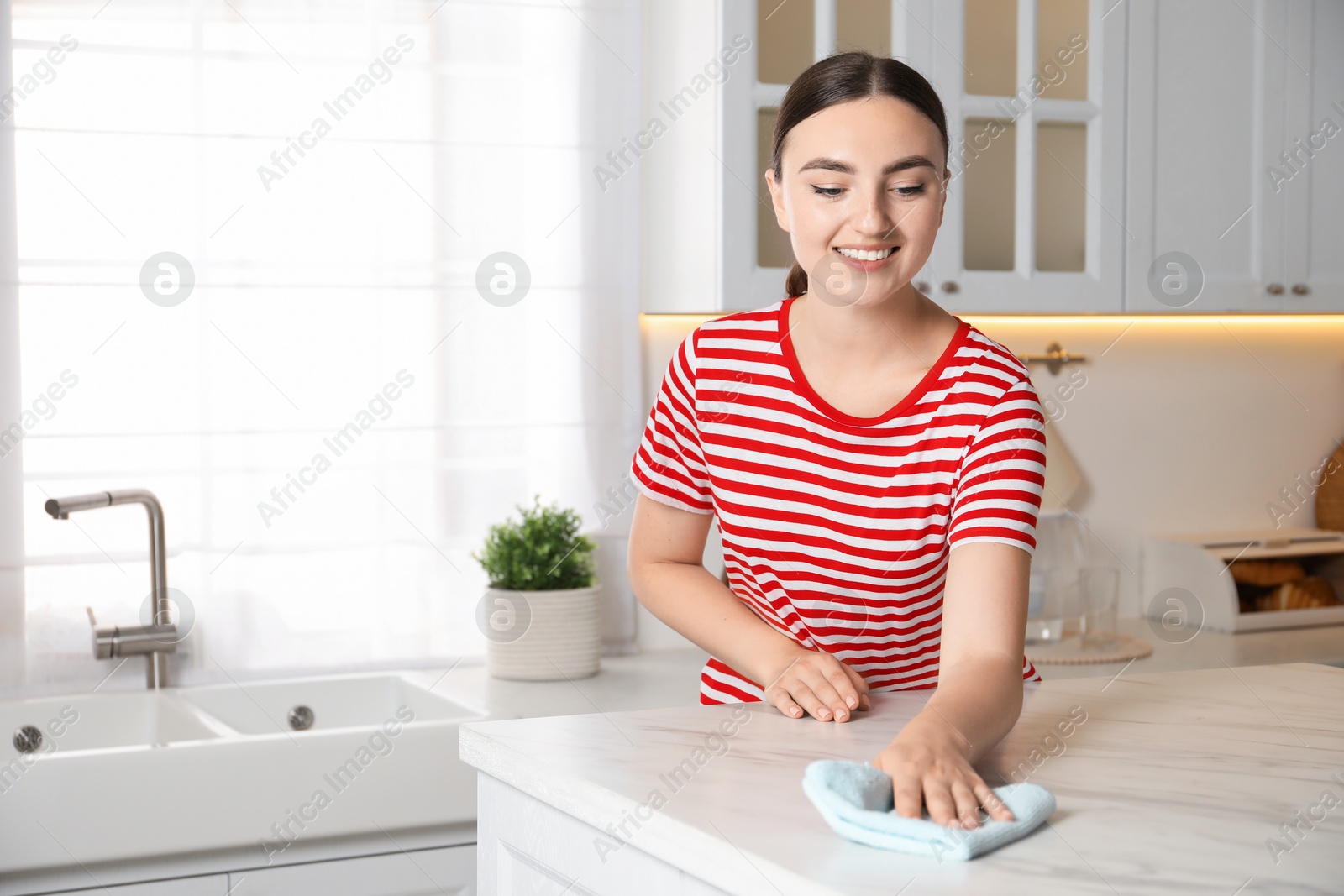 Photo of Beautiful young woman cleaning white marble table with rag in kitchen