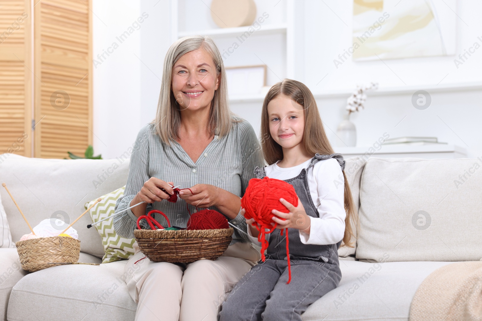 Photo of Happy grandmother with granddaughter holding knitting needles and yarn on sofa at home