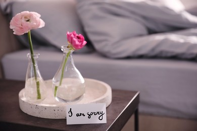 Image of Sorry card and flowers on bedside table in room. Space for text