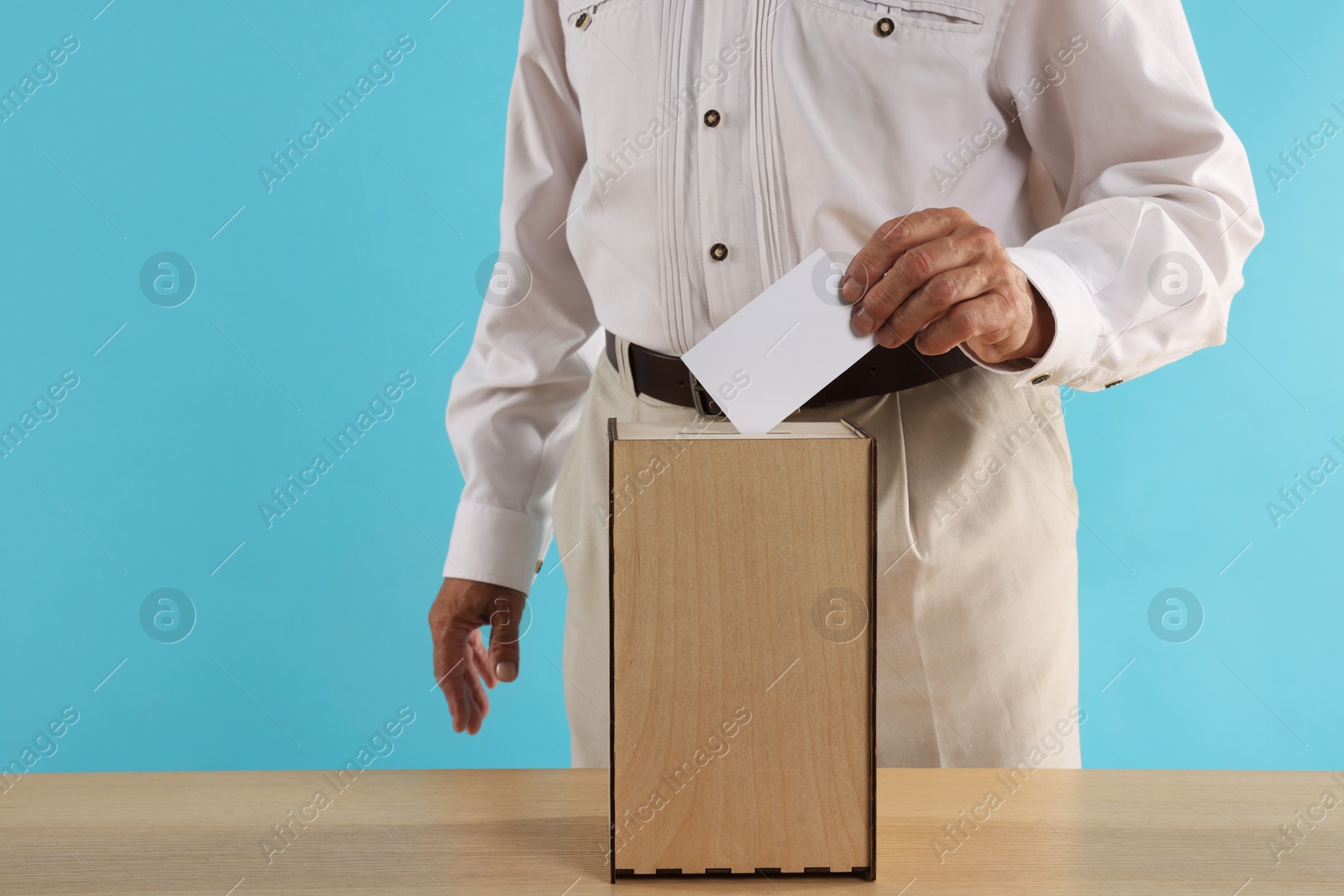 Photo of Referendum. Man putting his vote into ballot box at wooden table against light blue background, closeup