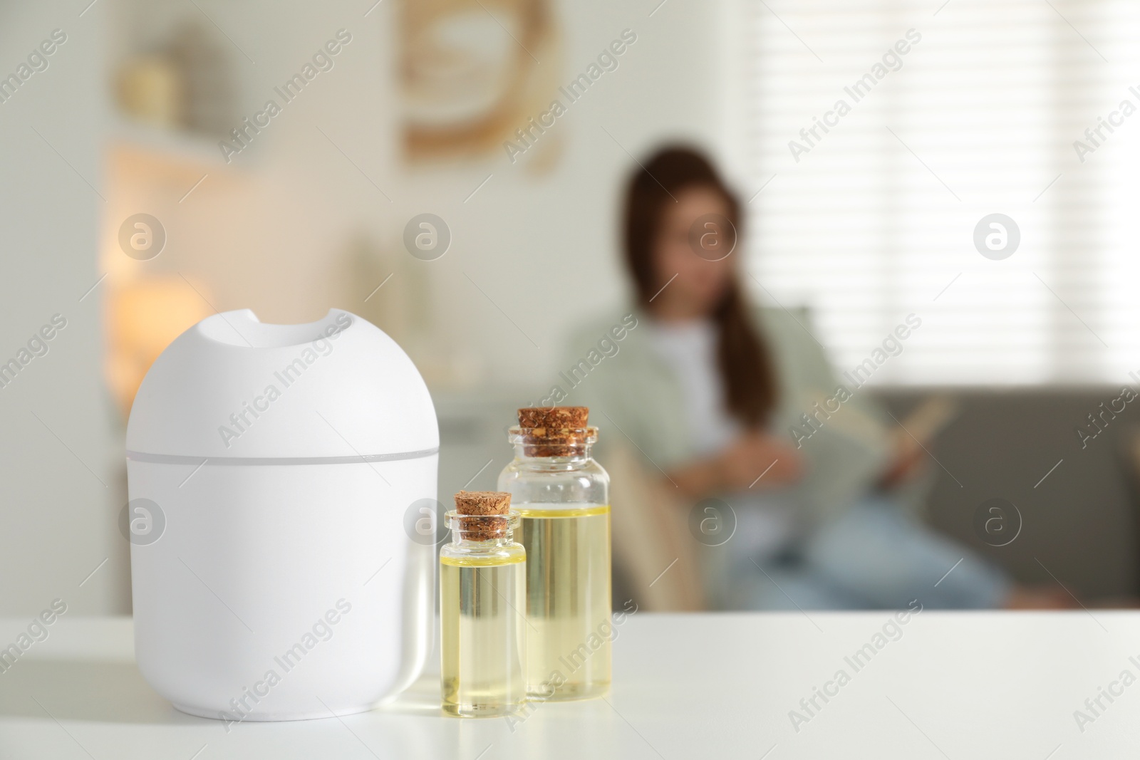 Photo of Woman reading book at home, focus on aroma diffuser and bottles of essential oils