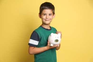 Photo of Pocket money. Cute boy with piggy bank and coins on yellow background