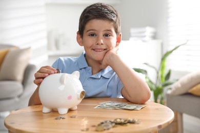 Pocket money. Cute boy putting coin into piggy bank at wooden table indoors