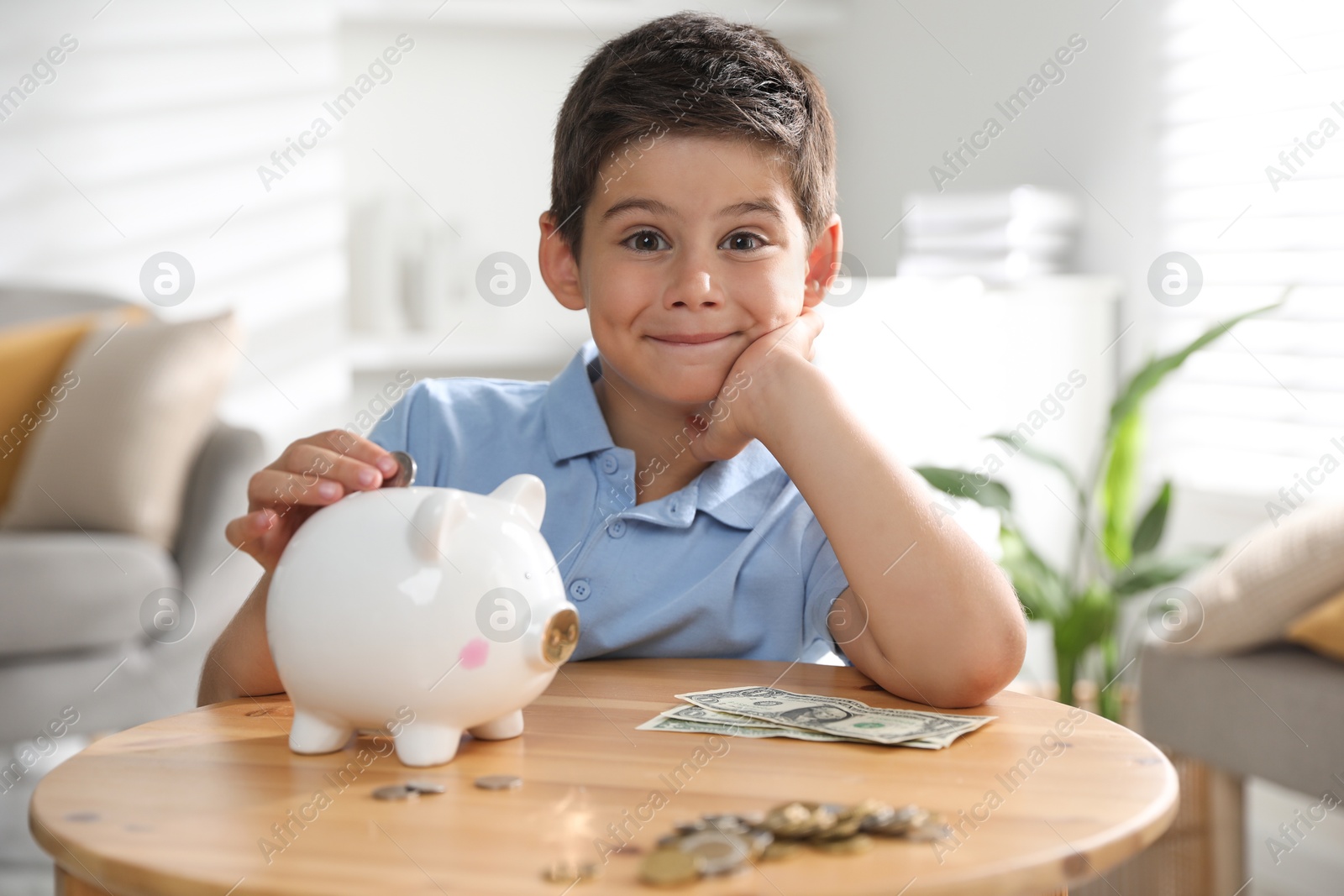 Photo of Pocket money. Cute boy putting coin into piggy bank at wooden table indoors