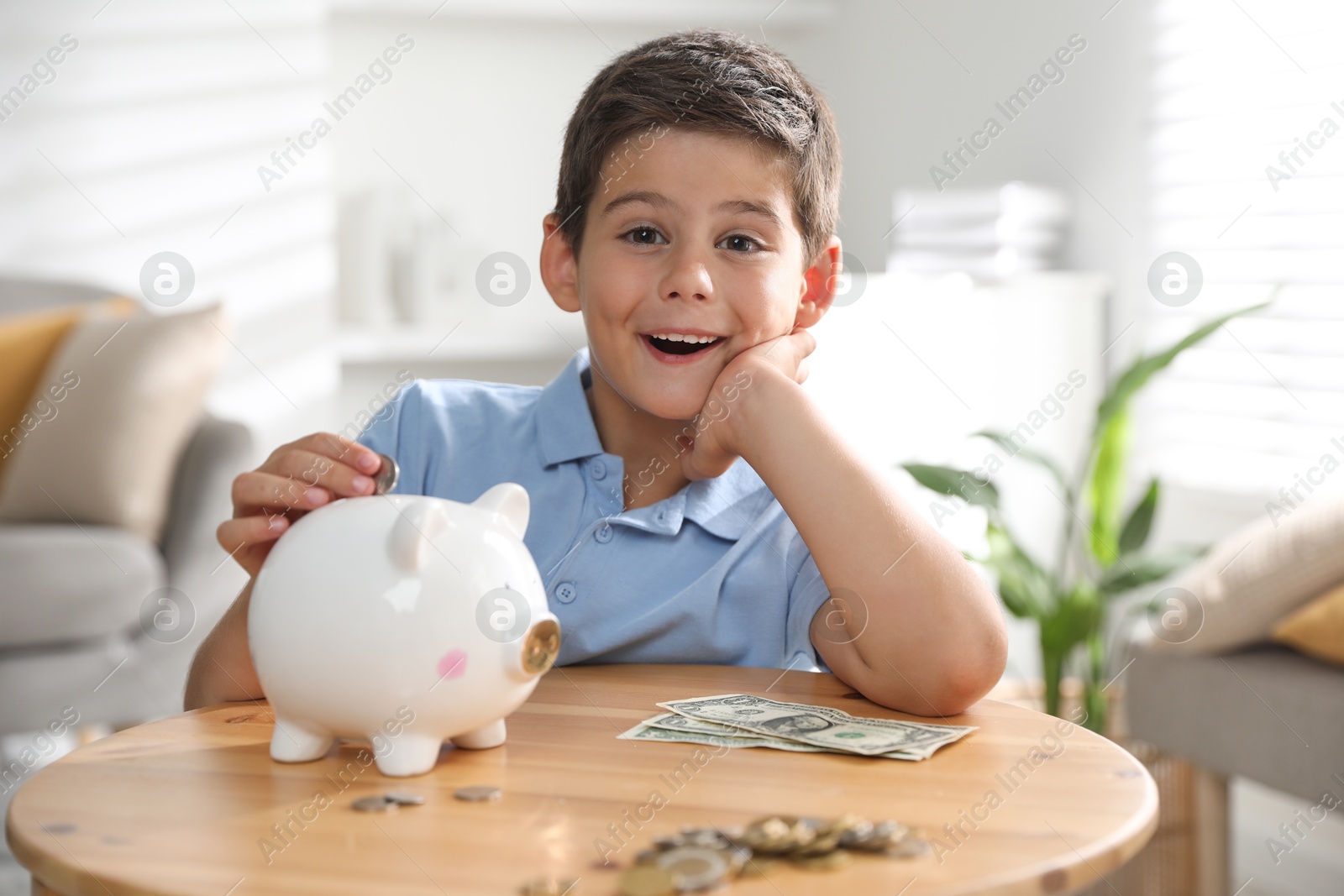 Photo of Pocket money. Cute boy putting coin into piggy bank at wooden table indoors