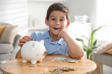 Pocket money. Cute boy putting coin into piggy bank at wooden table indoors