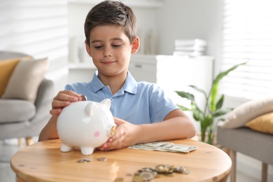 Photo of Pocket money. Cute boy putting coin into piggy bank at wooden table indoors