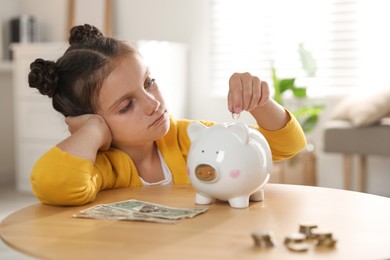 Photo of Pocket money. Cute girl putting coin into piggy bank at wooden table indoors