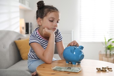 Pocket money. Cute girl putting coin into piggy bank at wooden table indoors