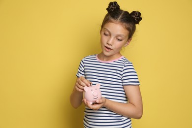 Photo of Pocket money. Cute girl putting coin into piggy bank on yellow background