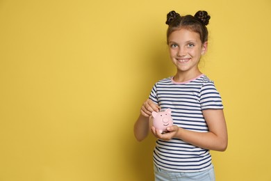 Photo of Pocket money. Cute girl putting coin into piggy bank on yellow background, space for text