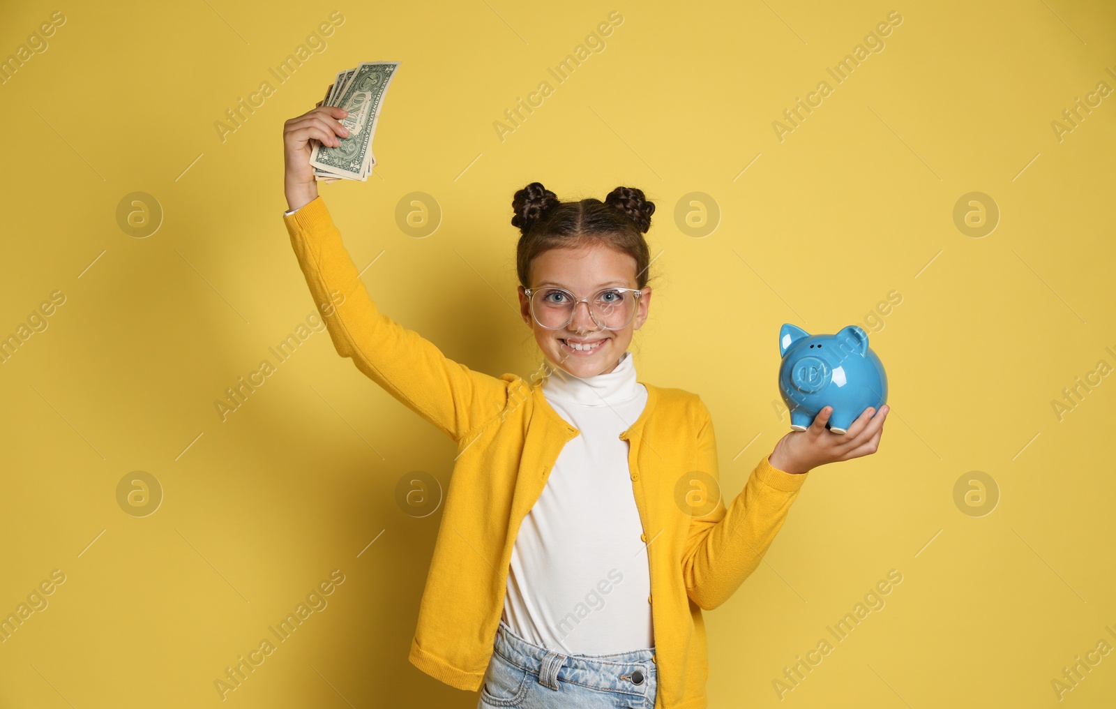 Photo of Pocket money. Cute girl with piggy bank and dollar banknotes on yellow background