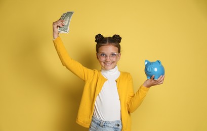 Photo of Pocket money. Cute girl with piggy bank and dollar banknotes on yellow background