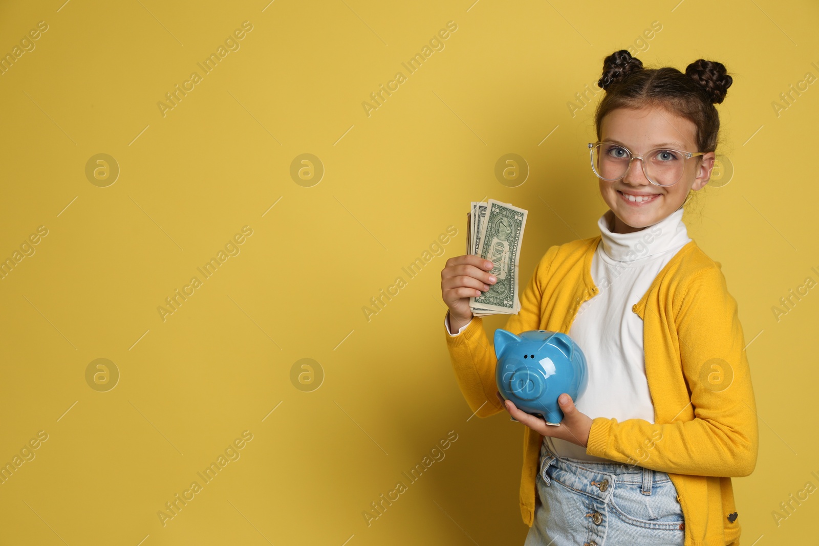 Photo of Pocket money. Cute girl with piggy bank and dollar banknotes on yellow background, space for text