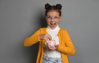 Pocket money. Cute girl putting coins into piggy bank on grey background