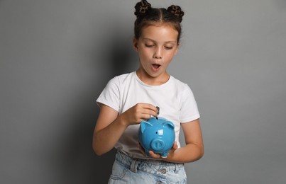 Photo of Pocket money. Cute girl putting coins into piggy bank on grey background