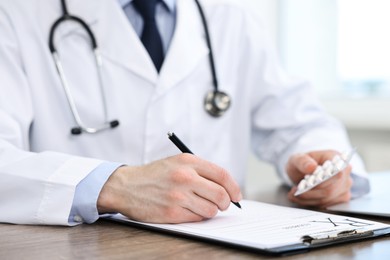 Photo of Doctor with pills writing prescription at wooden table in clinic, closeup