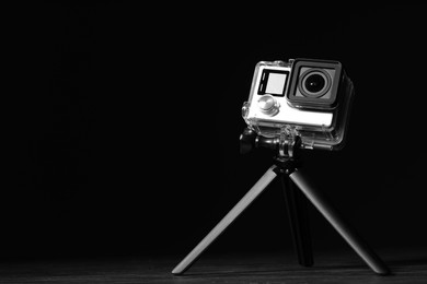 Modern action camera and tripod on wooden surface against black background. Space for text