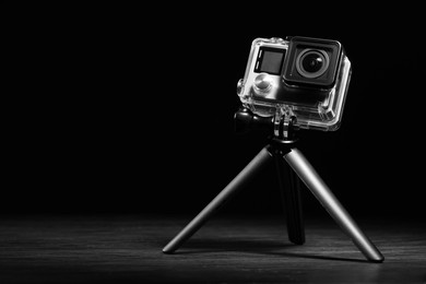 Modern action camera and tripod on wooden surface against black background. Space for text