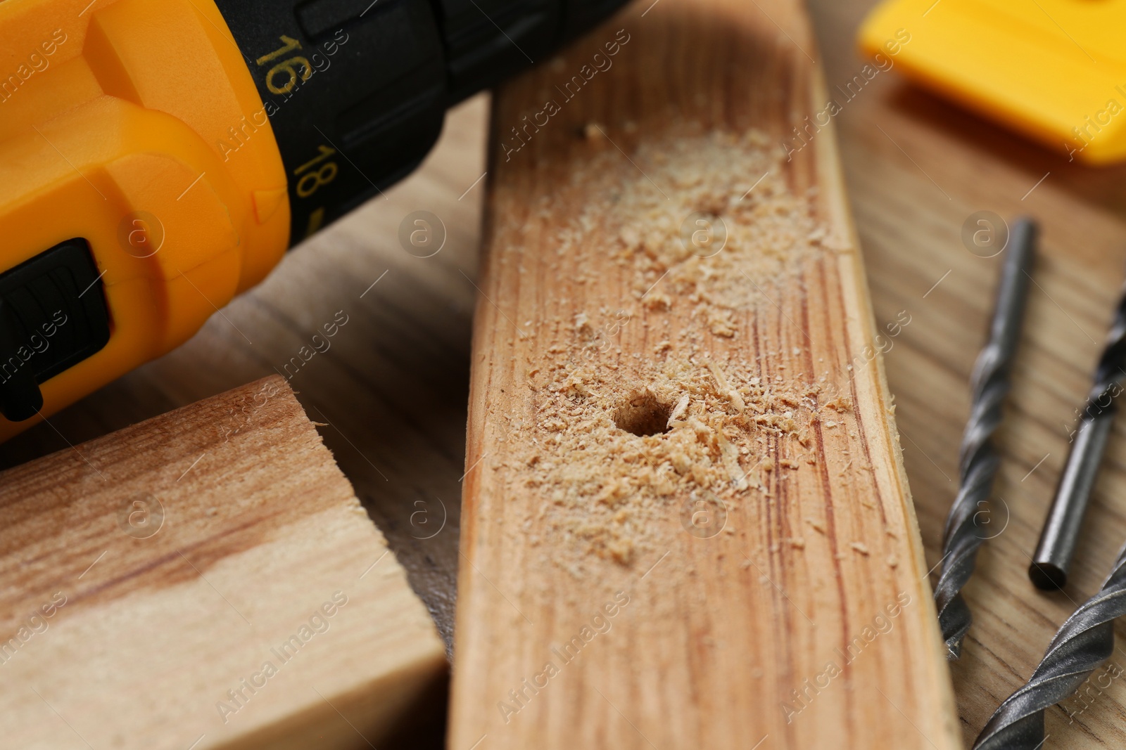 Photo of Cordless electric drill, bits, holed plank and sawdust on wooden table, closeup