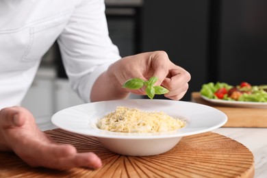 Photo of Professional chef decorating delicious pasta with basil at table in kitchen, closeup