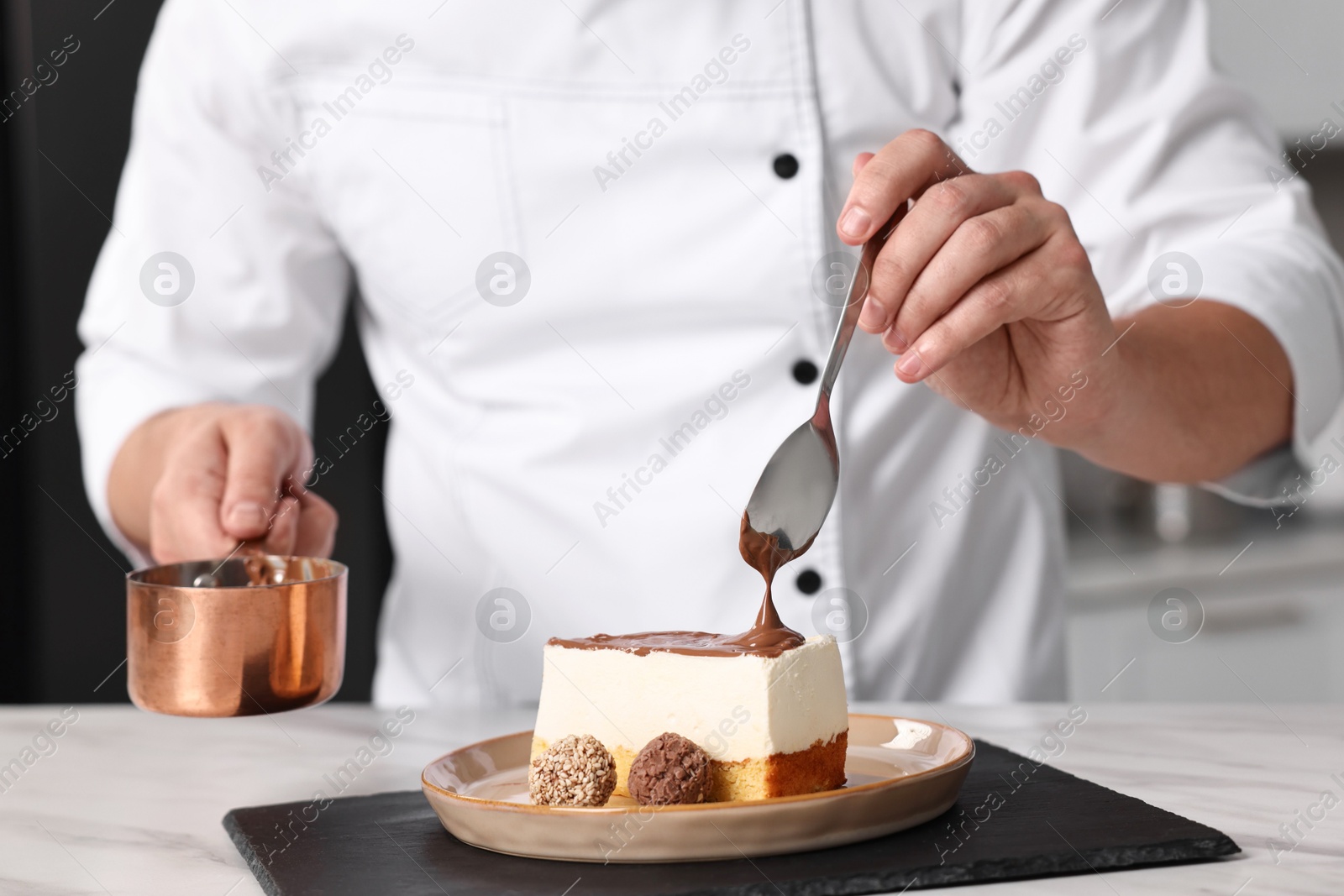 Photo of Professional chef pouring chocolate cream onto delicious cake at white marble table in kitchen, closeup