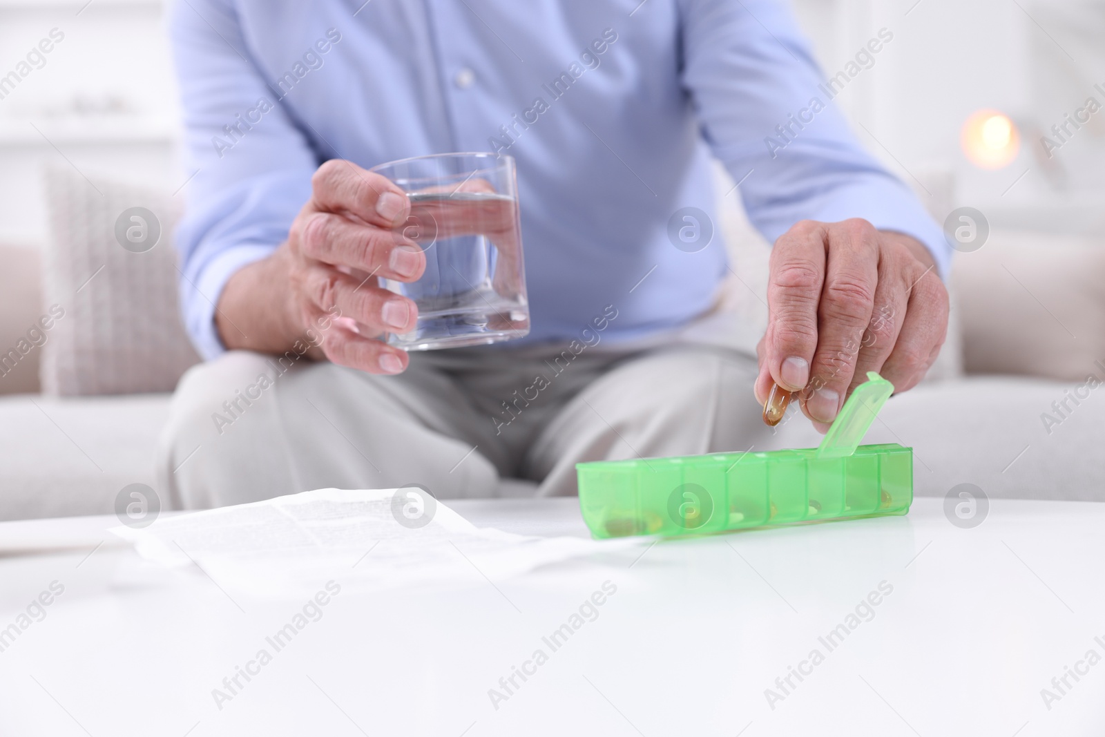 Photo of Senior man with glass of water taking pill from organizer at home, closeup