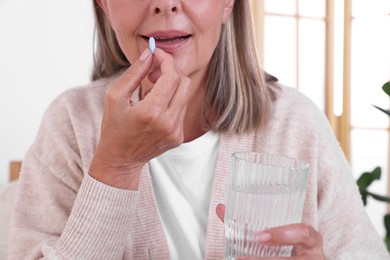 Photo of Senior woman with glass of water taking pill at home, closeup