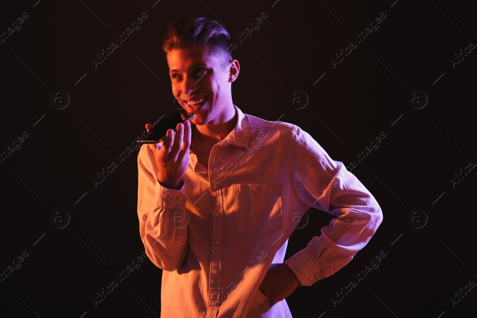 Photo of Young man recording voice message via smartphone on dark background with neon lights