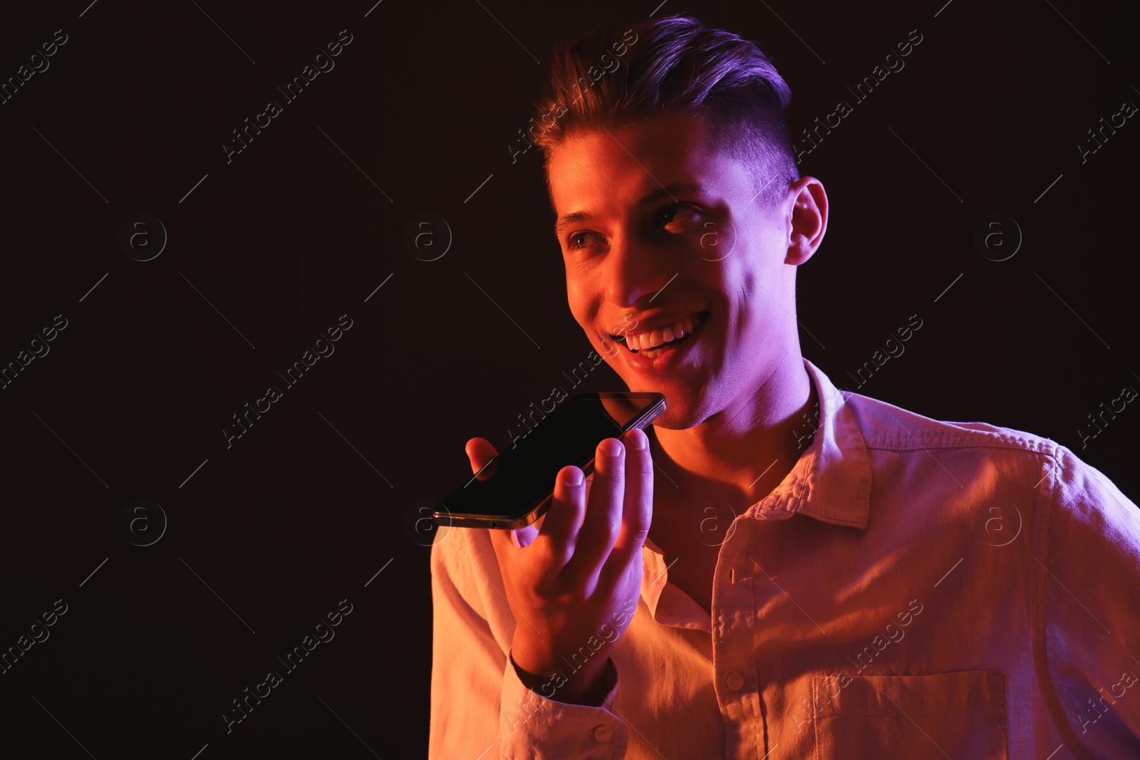 Photo of Young man recording voice message via smartphone on dark background with neon lights, space for text