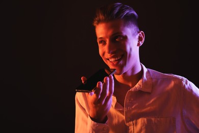 Young man recording voice message via smartphone on dark background with neon lights, space for text