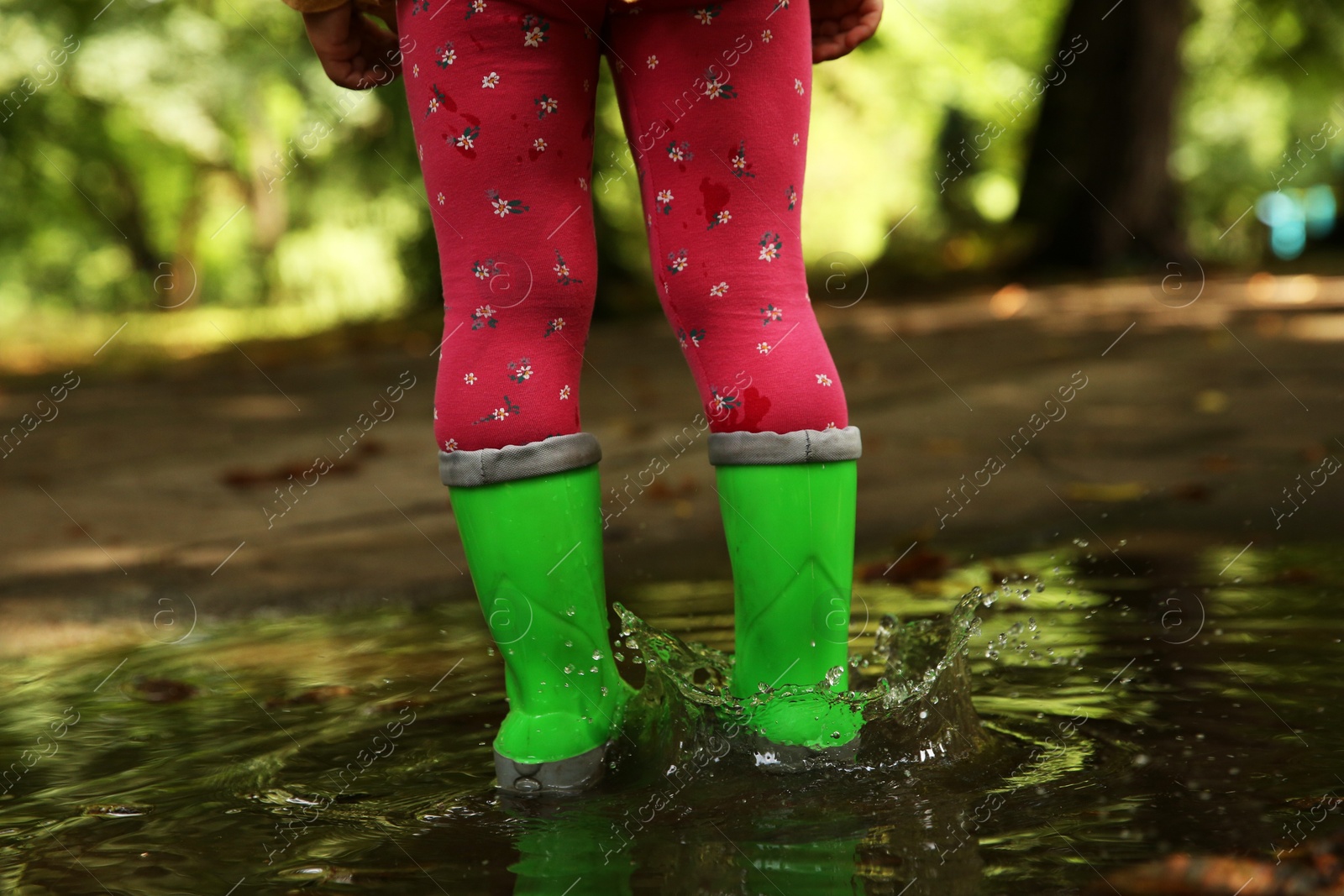 Photo of Little girl wearing green rubber boots standing in puddle outdoors, closeup