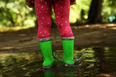 Photo of Little girl wearing green rubber boots standing in puddle outdoors, closeup