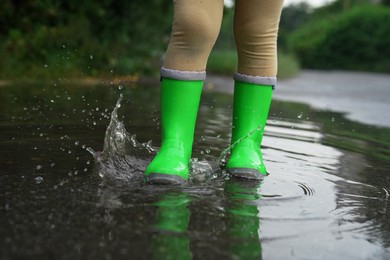 Photo of Little girl wearing green rubber boots walking in puddle outdoors, closeup