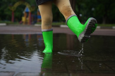 Photo of Little girl wearing green rubber boots walking in puddle outdoors, closeup