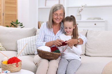 Photo of Smiling grandmother teaching her granddaughter to knit on sofa at home