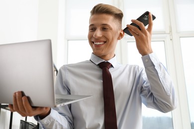 Photo of Young man with laptop listening to voice message via smartphone in office